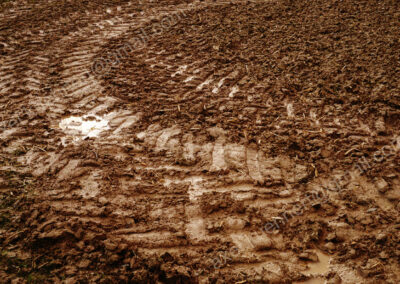 In einen durchgeweichten satt-braunem Ackerboden haben im Bogen fahrende Traktoren pfeilförmige Reifenmuster eingeprägt,